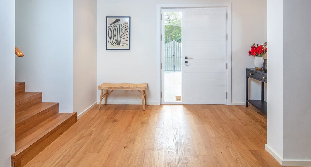 A bright entryway in a Heritage villa with oak wood flooring, a white front door, staircase, black cabinet, and a small bench against the wall.