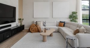 A living room in Sanctuary Falls featuring modern flooring materials and a grey carpet, with a grey sofa, pillows, light brown coffee table, wall-mounted TV, and black TV bench.