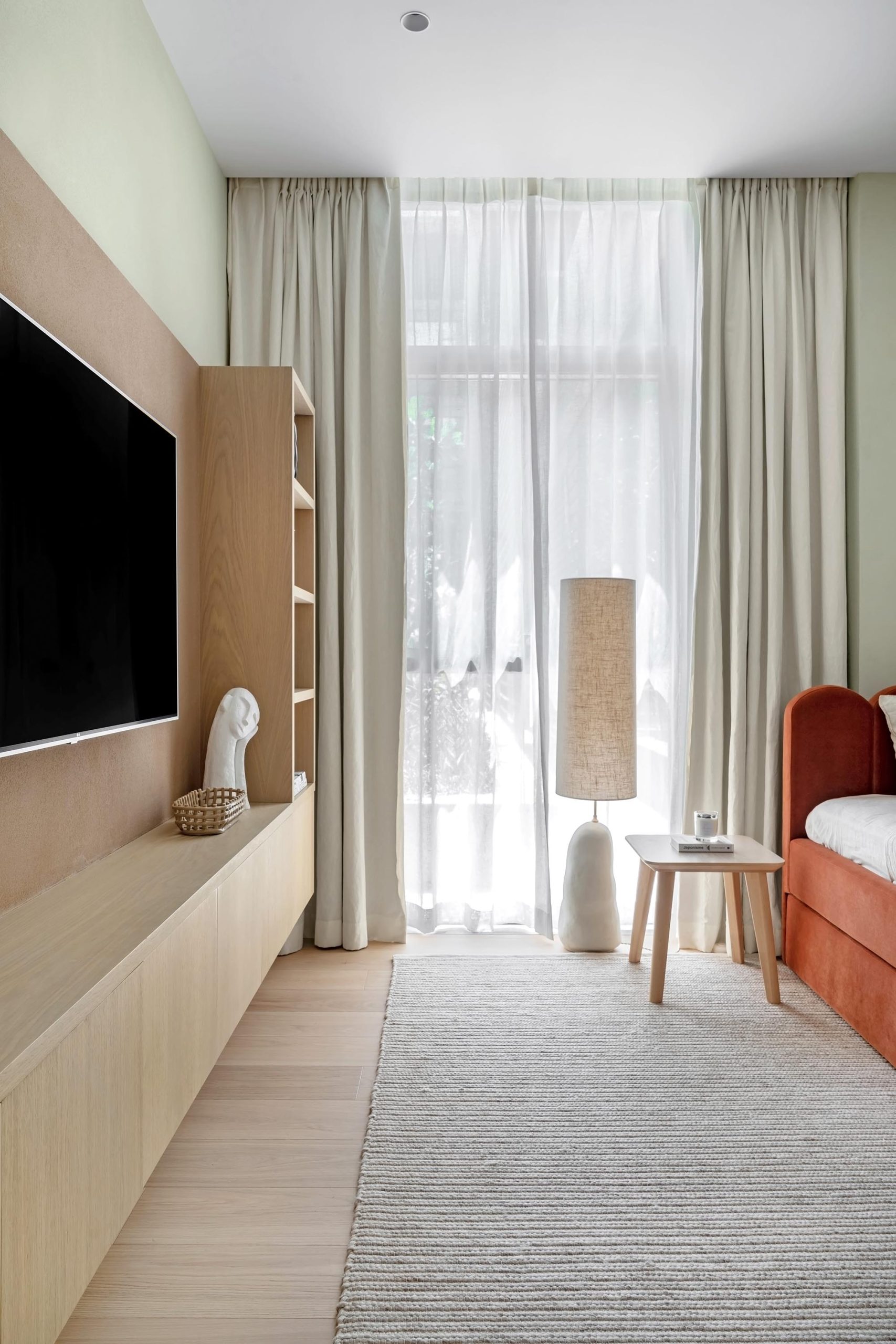 A minimalist living room with a light wood flooring design, a wall-mounted TV, sheer curtains, and a red-orange sofa.