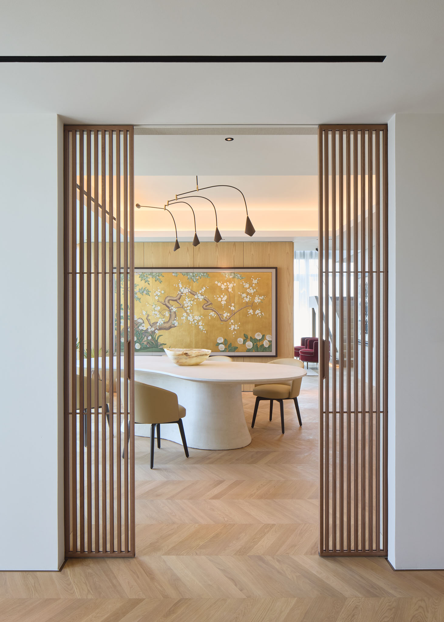 A dining room with a curved dining table, beige chairs, a light Chevron wood flooring design, and wooden slatted partitions.