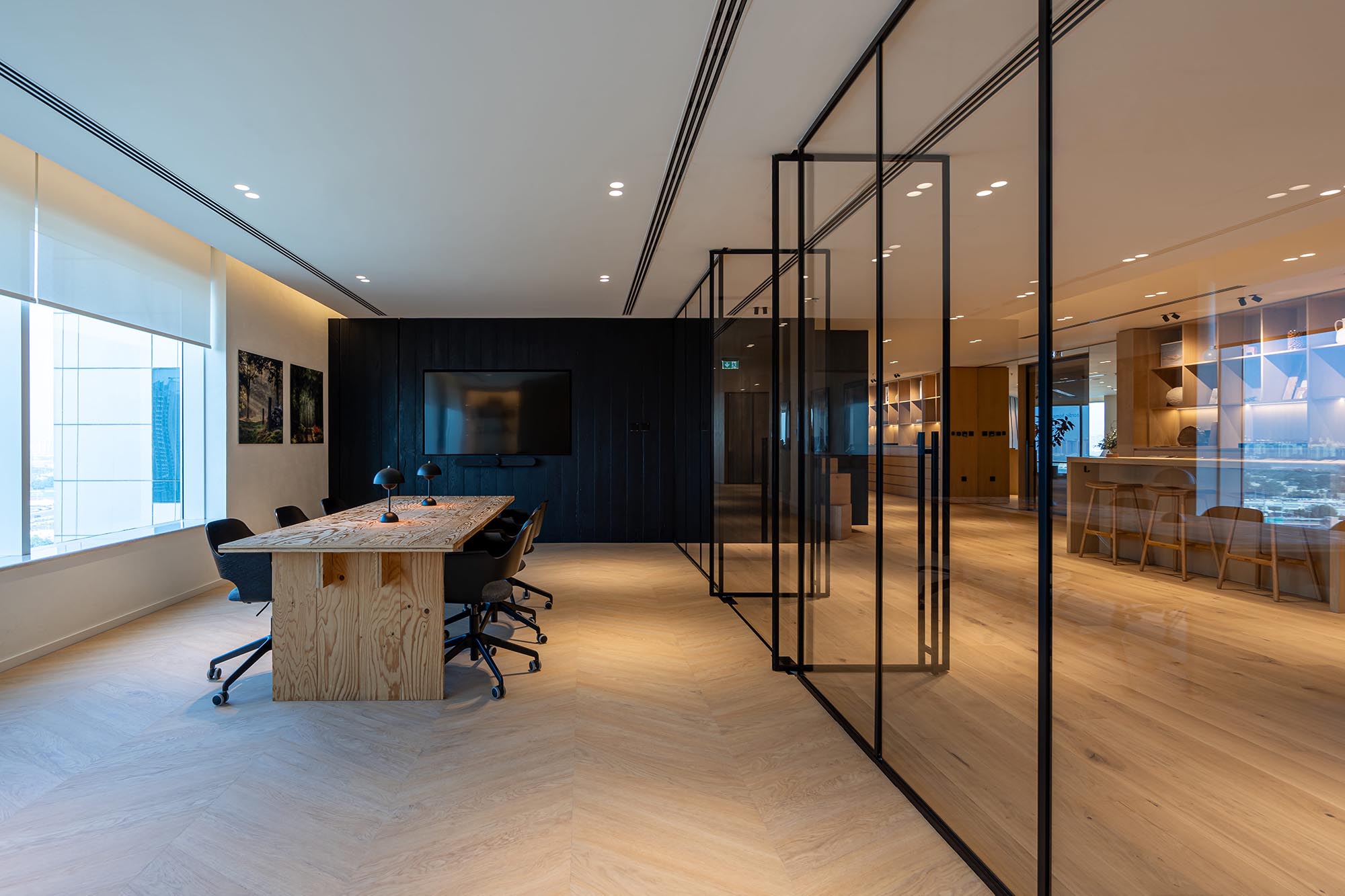  A modern office meeting room with light Chevron wood flooring design, a large conference table, black chairs, and glass partition walls. 