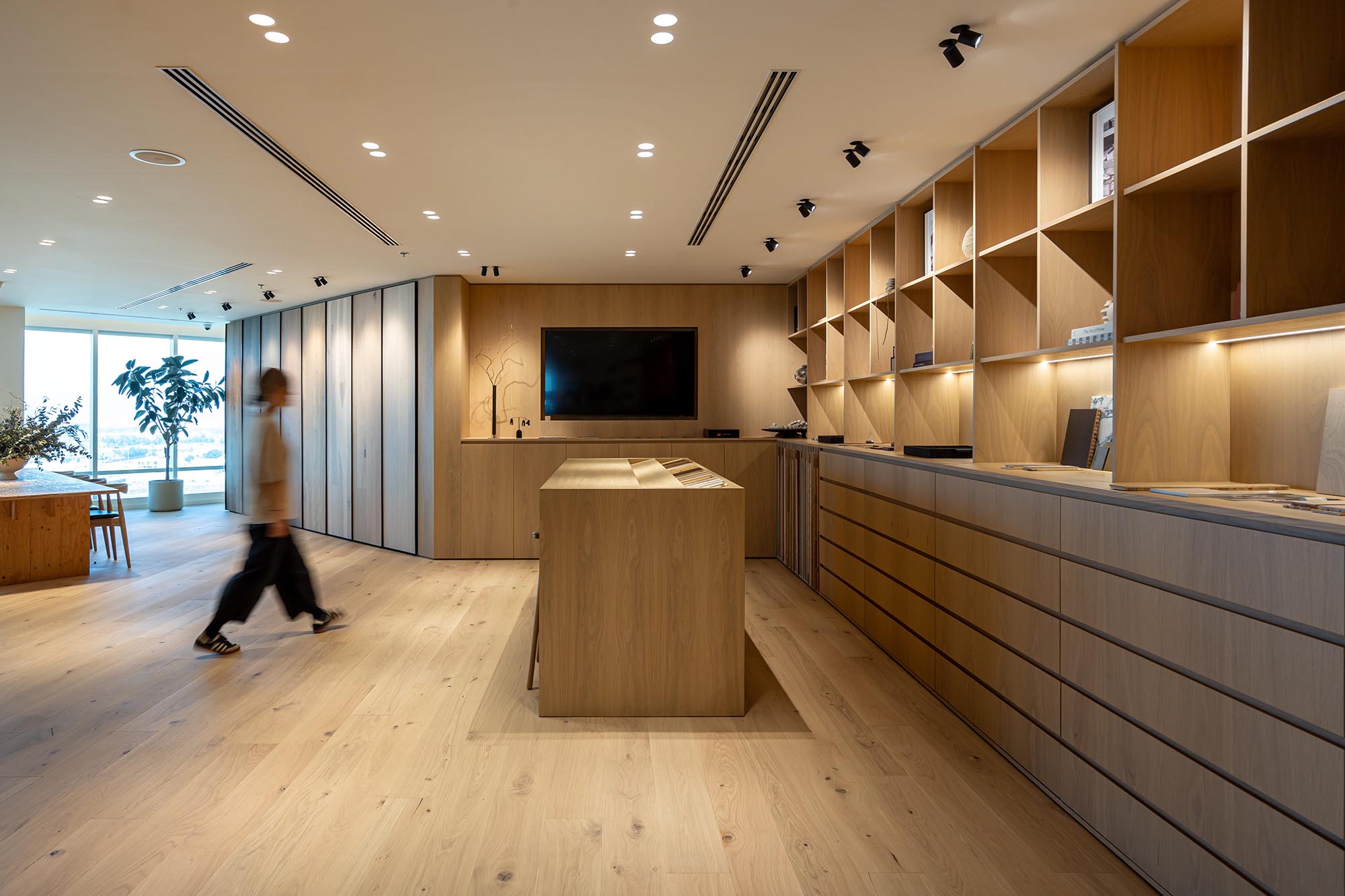 A dining and showcase room with light wooden flooring designs, an island with chairs, a wall-mounted TV, wooden cabinetry, and a woman walking in between