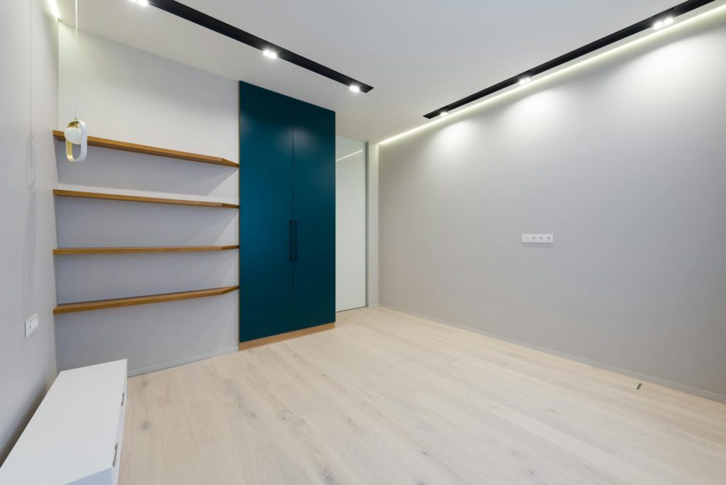 A room with laminate flooring, grey walls, brown wall shelves, and a dark blue cabinet, showcasing types of flooring.
