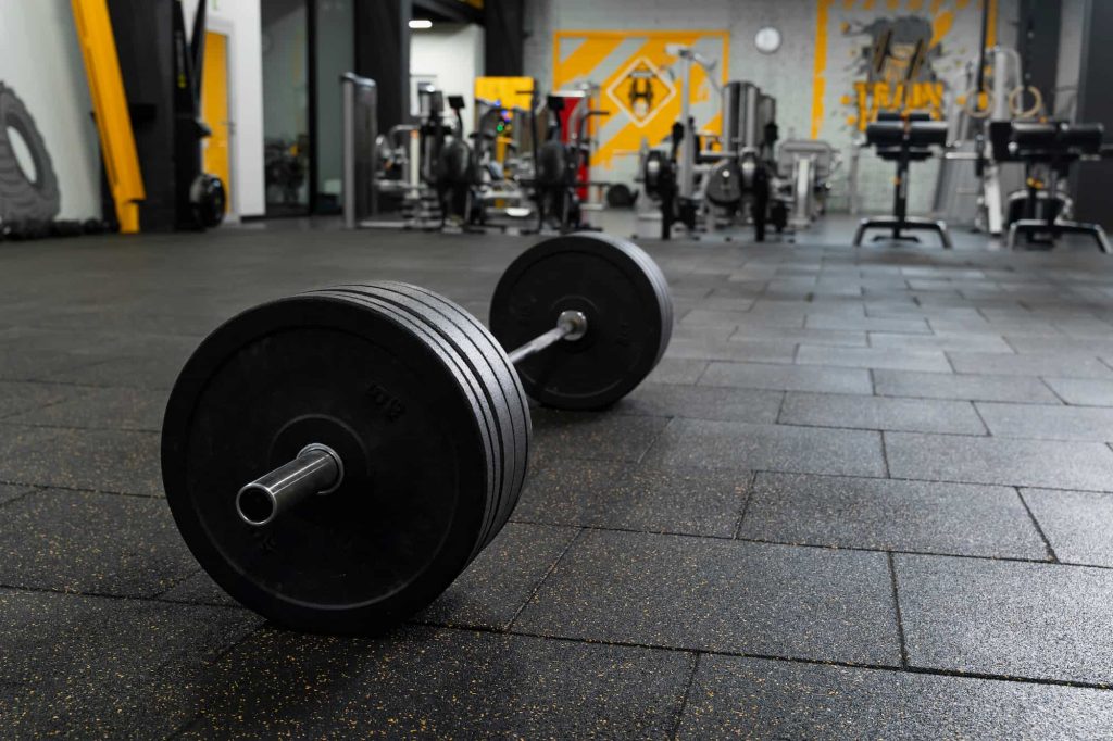 Black rubber flooring in a gym with a barbell resting on the floor. The background is full of gym equipment.